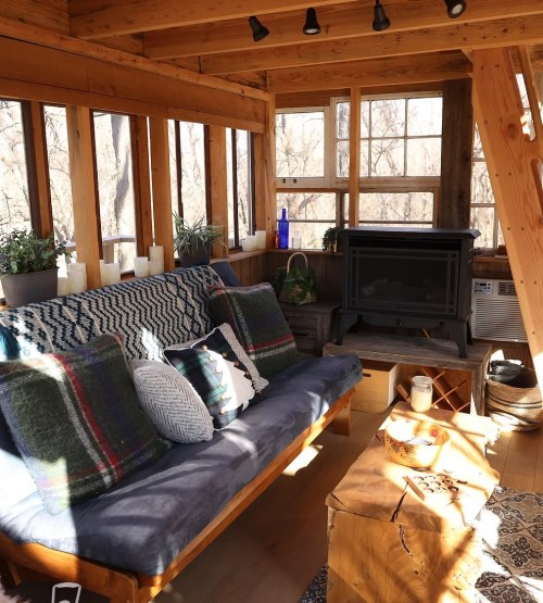 Cozy interior of a wooden cabin with a sofa, plants, and a wood stove, surrounded by large windows.