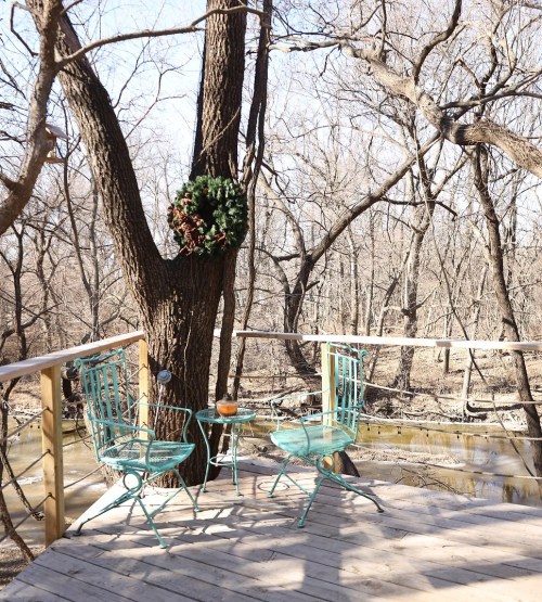 A cozy outdoor seating area with two teal chairs and a small table, surrounded by bare trees and a river.