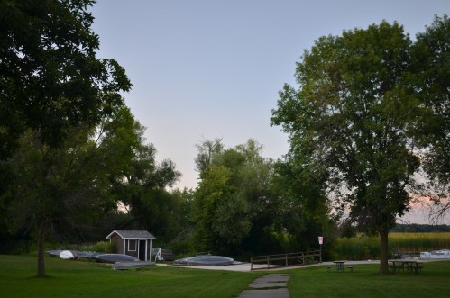 A serene park scene with trees, a small shed, and covered boats near a grassy path leading to a calm waterway.