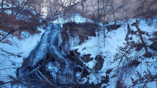 A frozen waterfall cascading down a snowy cliff, surrounded by bare trees and rocky terrain.