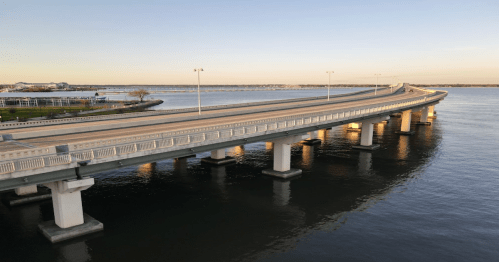 A bridge spans over calm water, with streetlights illuminating the roadway at sunset.