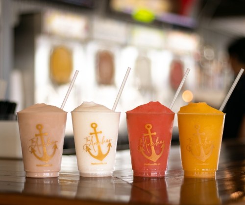 Four colorful frozen drinks in clear cups with straws, displayed on a wooden counter.