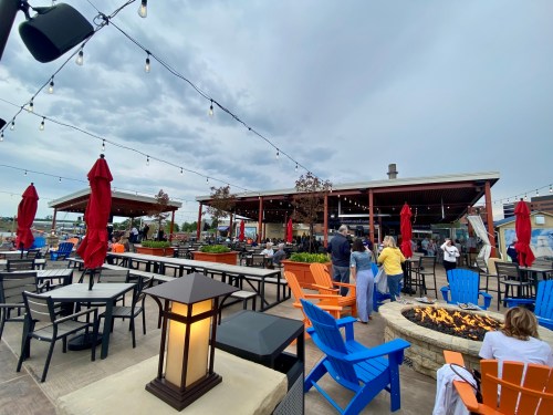 Outdoor dining area with colorful chairs, string lights, and a fire pit, set against a cloudy sky.