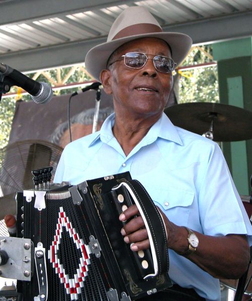 A man in a light blue shirt and hat plays an accordion on stage, smiling at the audience.