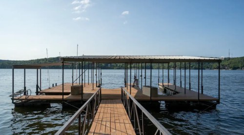 A wooden dock extends over calm water, surrounded by green hills under a clear blue sky.