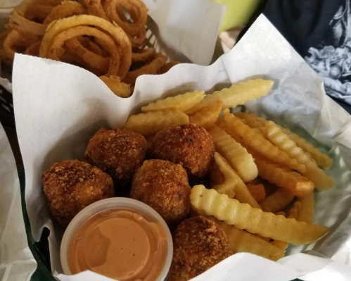 A basket of crispy fried food including onion rings, crinkle-cut fries, and golden-brown balls with dipping sauce.