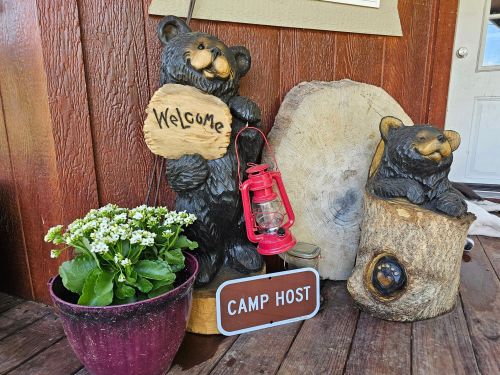 Two bear sculptures with a "Welcome" sign, a red lantern, a plant, and a "Camp Host" sign on a wooden porch.