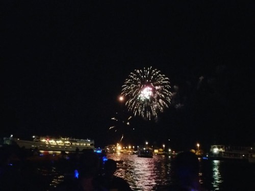 Colorful fireworks illuminate the night sky over a waterfront, with boats and city lights in the foreground.