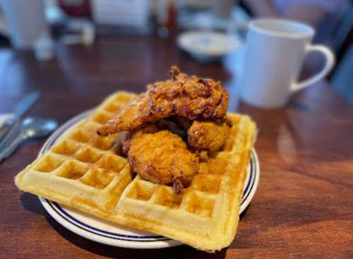 A plate of waffles topped with fried chicken pieces, served on a wooden table with a coffee cup in the background.