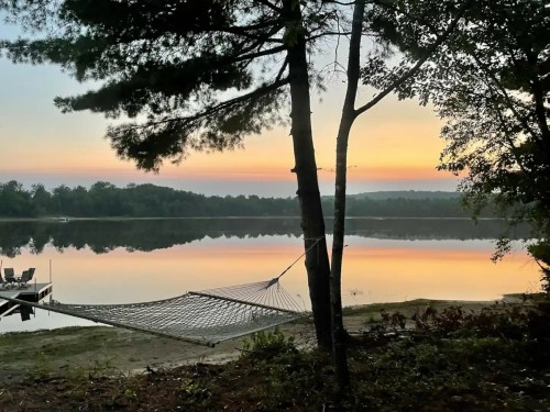 A serene lake at sunset, with a hammock hanging between trees and a calm reflection on the water.