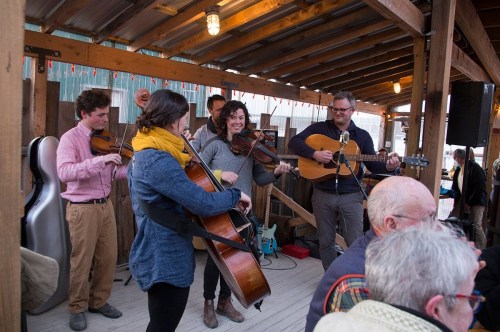 A lively band performs with violins and a guitar in a rustic venue, while an audience enjoys the music.