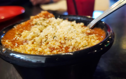 A close-up of a bowl of chili topped with cheese and herbs, with a spoon resting beside it on a dark table.