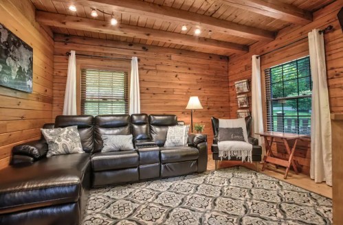 Cozy wooden living room with leather seating, a lamp, and a patterned rug, featuring large windows and warm lighting.