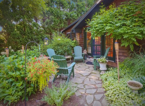 A cozy garden with stone pathways, green chairs, vibrant flowers, and a charming cottage in the background.