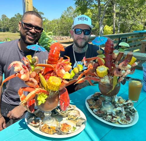 Two men sit at a table with large seafood cocktails, featuring shrimp, crab, corn, and garnishes, alongside a platter of oysters.