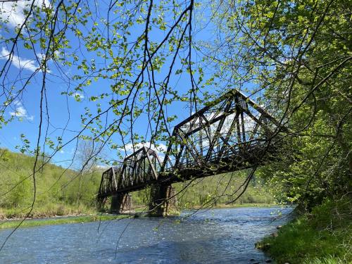 A rusted train bridge spans a river, surrounded by lush greenery and a clear blue sky.