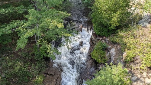 Aerial view of a flowing waterfall surrounded by lush green trees and rocky terrain.