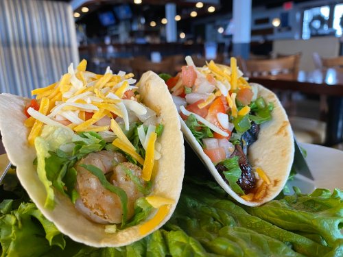 Two tacos filled with shrimp, lettuce, cheese, and fresh toppings, served on a bed of lettuce in a restaurant setting.