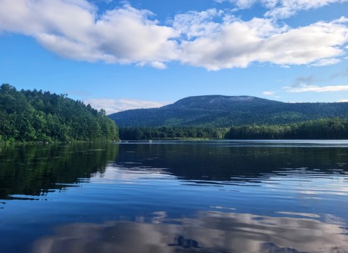A serene lake reflects a blue sky and clouds, surrounded by lush green trees and a distant mountain.