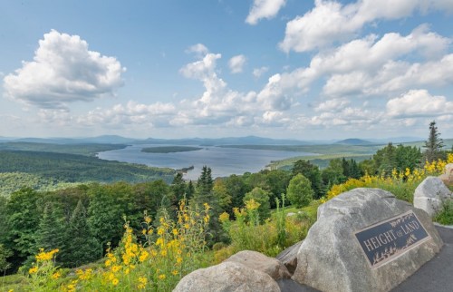 Scenic view from Height of Land, showcasing a lake surrounded by lush forests and mountains under a cloudy sky.