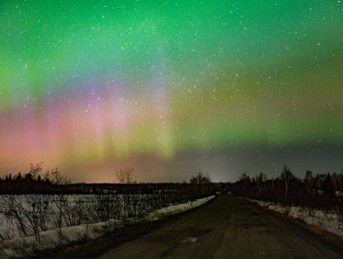 A serene road under a vibrant aurora borealis, with green and purple hues lighting up the night sky.