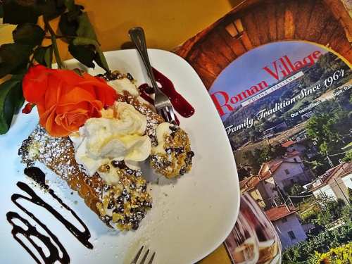 A plate of cannoli topped with whipped cream and a rose, next to a menu for Roman Village restaurant.