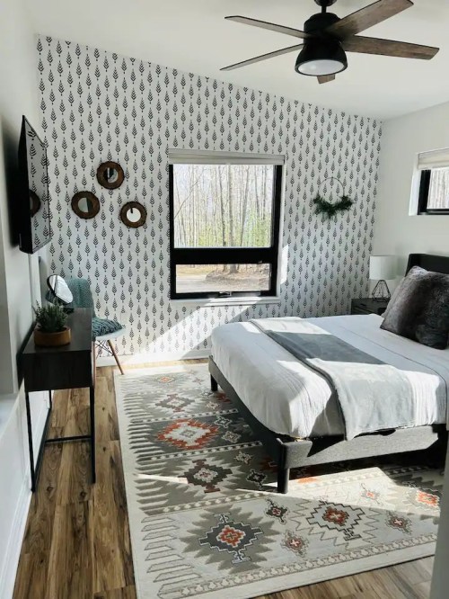 Cozy bedroom with patterned wallpaper, a bed, desk, and large window overlooking a forested area.