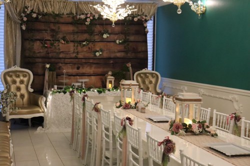 Elegant wedding reception setup with a long table, gold accents, floral decor, and a rustic wooden backdrop.