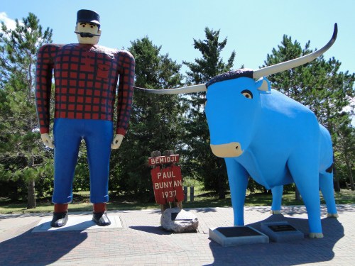 A large statue of Paul Bunyan in plaid and a blue ox stand together in a park, with trees in the background.
