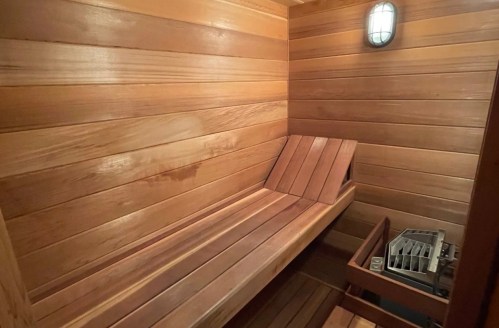 A wooden sauna interior featuring bench seating and a small heater, illuminated by a wall-mounted light.