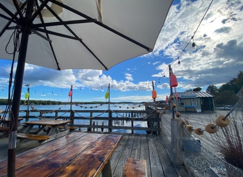 A wooden deck with a table and umbrella overlooks a calm waterway, decorated with colorful buoys and a cloudy sky.