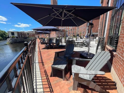 Outdoor seating area with gray chairs and umbrellas by a river, surrounded by brick buildings and blue sky.
