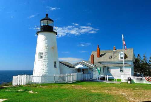 A white lighthouse beside a coastal house, with a blue sky and ocean in the background, surrounded by green grass.