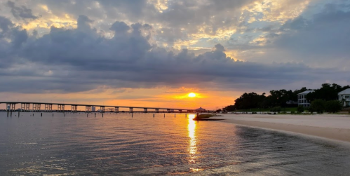 Sunset over calm waters with a bridge in the background and a sandy beach lined with trees and houses.