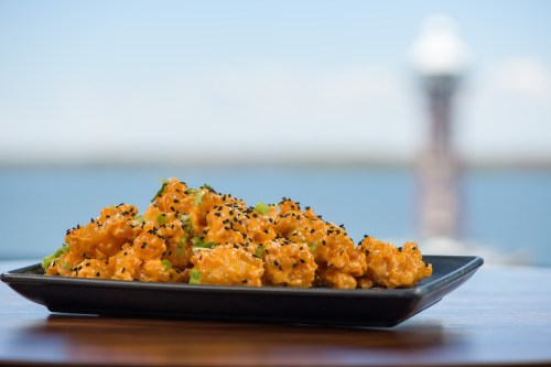 A plate of orange-coated, sesame-topped shrimp with green garnish, set against a blurred waterfront background.