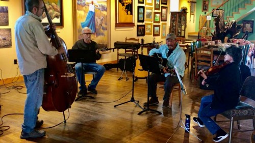 A group of four musicians play instruments in a cozy, art-filled space, creating a lively atmosphere.