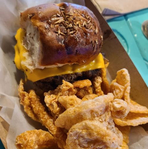 A cheeseburger with a soft bun and crispy pork rinds on the side, served in a paper tray.