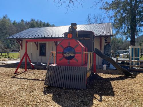 A colorful train-themed playground structure with a slide, swings, and a building in the background surrounded by trees.