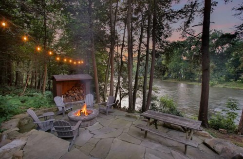 Cozy outdoor fire pit area by a river, surrounded by trees and string lights, creating a serene atmosphere.