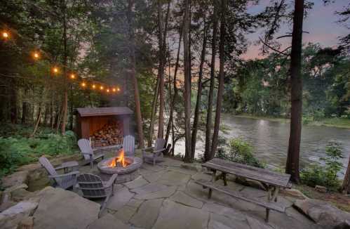 Cozy outdoor fire pit area by a river, surrounded by trees and string lights, creating a serene atmosphere.