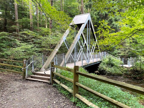 A metal bridge with a triangular design spans a small creek, surrounded by lush green trees and a wooden fence.