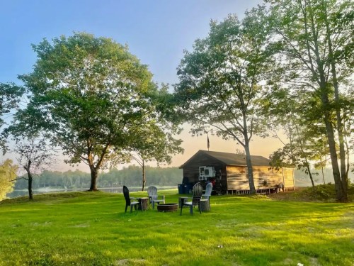 A cozy cabin surrounded by trees, with chairs arranged around a fire pit on a grassy area by a lake.