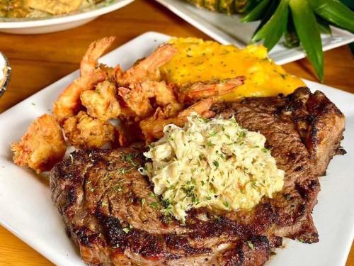 A plate featuring a grilled steak topped with crab meat, accompanied by fried shrimp and a side of cheesy grits.