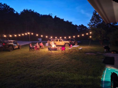 A cozy outdoor gathering around a campfire, with string lights, vehicles, and people sitting in chairs.