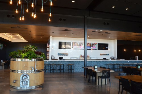 Modern bar area with a wooden barrel planter, sleek counter, and multiple screens displaying content.