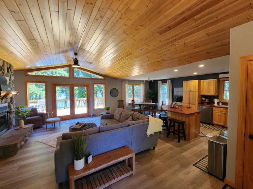Cozy living room with wooden ceiling, large windows, and open kitchen, featuring a comfortable sofa and dining area.