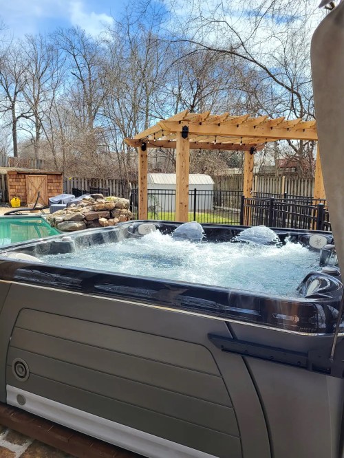 A hot tub with bubbling water, surrounded by trees and a wooden pergola in a backyard setting.