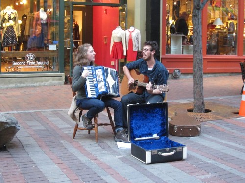 A man plays guitar while a woman plays accordion, both seated on a street with shops in the background.