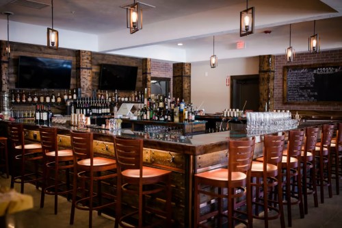A stylish bar with wooden decor, a long counter, and shelves filled with various bottles and glasses.
