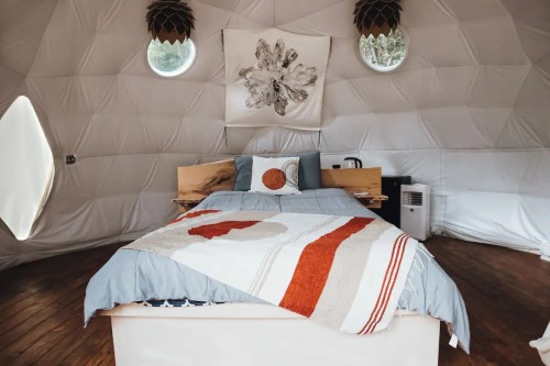 Cozy interior of a geodesic dome with a bed, decorative pillows, and natural light from circular windows.